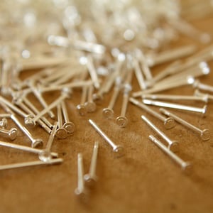 May include: A close-up of a pile of silver-colored metal pins with rounded heads. The pins are scattered on a brown surface.
