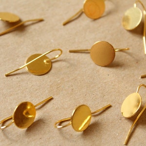 May include: A collection of gold-toned earring findings with round, flat, disc-shaped tops. The findings are scattered on a brown surface.