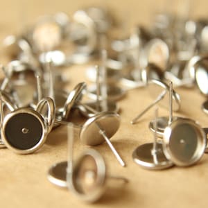 May include: A close-up of a group of silver earring settings. The settings are round and have a small hole in the center. They are scattered on a brown surface.