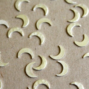 May include: A close-up of a group of silver crescent moon charms scattered on a brown surface. The charms are all the same size and shape, and they have a slightly textured finish.