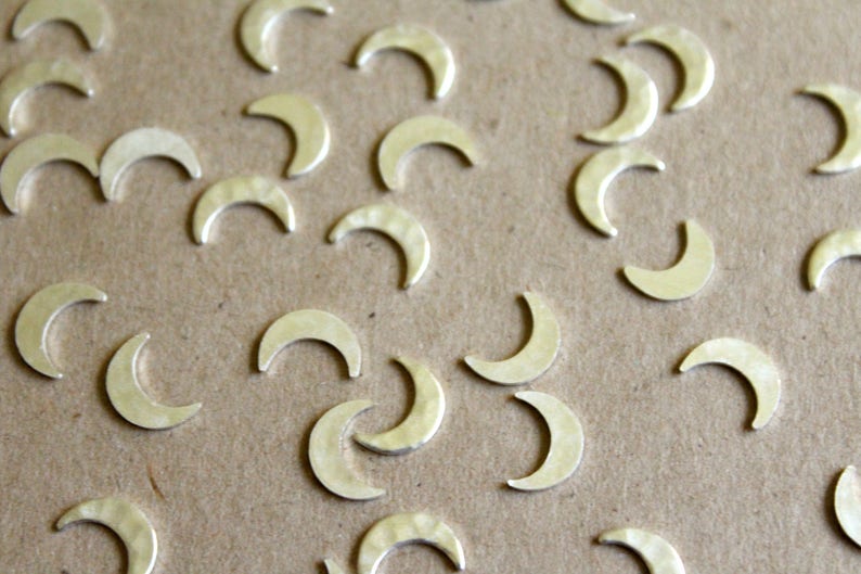 May include: A close-up of a group of silver crescent moon charms scattered on a brown surface. The charms are all the same size and shape, and they have a slightly textured finish.