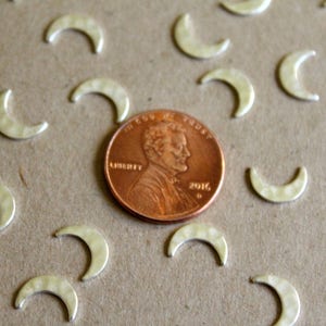 May include: A close-up of a group of silver crescent moon charms scattered on a brown surface. A penny is in the center of the image for scale.