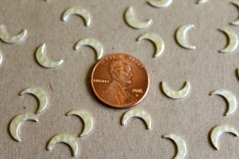 May include: A close-up of a group of silver crescent moon charms scattered on a brown surface. A penny is in the center of the image for scale.