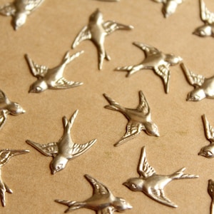 以下が含まれることがあります： 銀色の金属製のツバメのチャームのコレクション。チャームは茶色の表面に散らばっています。