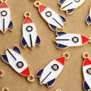 May include: A group of gold-coloured rocket ship charms with red, white and blue enamel paint. The charms have a loop at the top for attaching to jewellery or other crafts.