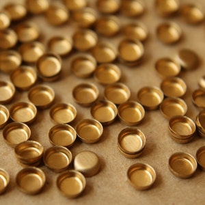 May include: A close-up of a large quantity of small, round, gold-colored metal caps. The caps are scattered on a brown surface.