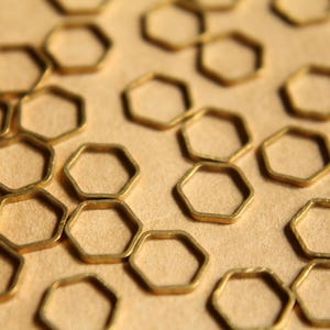 Könnte beinhalten: Nahaufnahme einer Gruppe goldfarbener sechseckiger Metallringe, die auf einer hellbraunen Oberfläche verstreut sind. Die Ringe sind alle gleich groß und gleich geformt und in einem zufälligen Muster angeordnet.