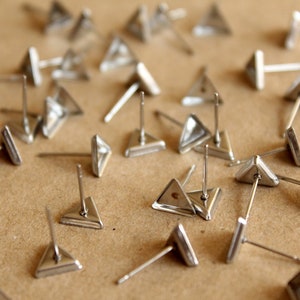 May include: A collection of silver triangle stud earrings scattered on a brown surface. The earrings have a simple, geometric design.