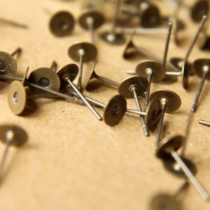 May include: A close-up of a pile of small, round, brass-colored metal tacks with sharp points. The tacks are scattered on a brown surface.