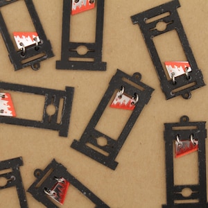 Könnte beinhalten: Eine Sammlung von Miniatur-Guillotine-Anhängern in Schwarz mit rot-weißem Klingendesign. Die Anhänger sind aus Metall gefertigt und haben oben eine kleine Schlaufe zum Befestigen an Schmuck oder anderen Bastelarbeiten. Der Hintergrund ist hellbraun.