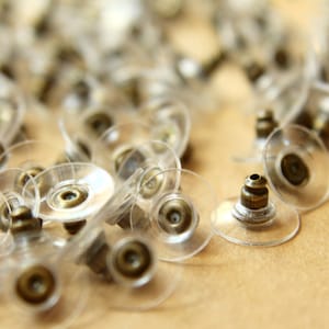 May include: A close-up of a pile of clear plastic earring backs with gold metal posts. The backs are designed to be used with stud earrings.