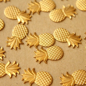 May include: A collection of gold-toned pineapple-shaped charms. The charms are arranged in a pattern on a brown surface.