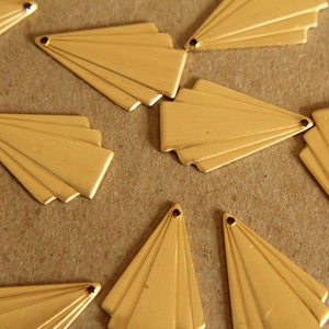 May include: Close-up of several gold-colored metal fan-shaped charms. Each charm has a small hole at the top for stringing. The charms are arranged on a brown surface. These charms are suitable for jewelry making.