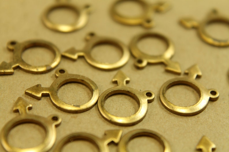 May include: A collection of gold-toned metal charms in the shape of a circle with an arrow pointing to the right. The charms are arranged on a brown surface.