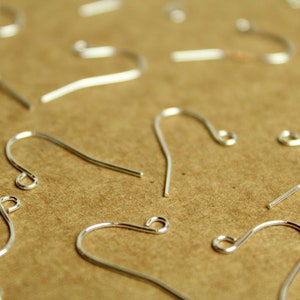 May include: Close-up of several silver-colored earring hooks. The hooks are made of thin, curved metal wire with a small loop at one end. The hooks are scattered on a light brown surface.