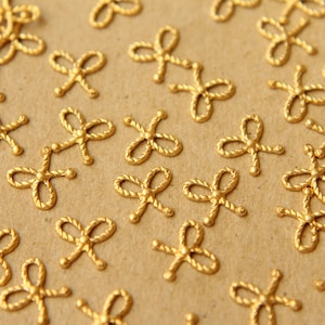 May include: A close-up of a group of gold-colored metal decorative embellishments in the shape of a bow. The embellishments are scattered on a brown surface.