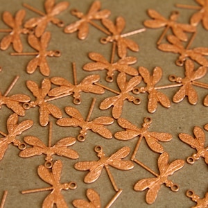 May include: A collection of small, copper-colored dragonfly charms. Each charm features detailed wings and a loop for attaching to jewelry or other crafts. The charms are scattered on a brown surface.