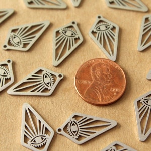 Op de afbeelding: Een close-up van verschillende zilveren metalen bedels in de vorm van een diamant met een oog- en zonnestralenontwerp. De bedels zijn verspreid over een bruin oppervlak naast een Amerikaanse penny.