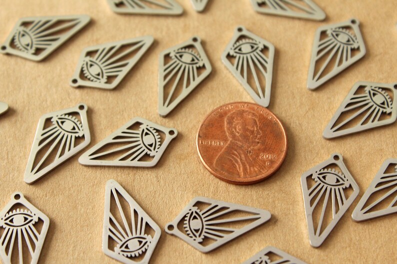 Op de afbeelding: Een close-up van verschillende zilveren metalen bedels in de vorm van een diamant met een oog- en zonnestralenontwerp. De bedels zijn verspreid over een bruin oppervlak naast een Amerikaanse penny.