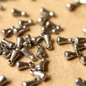 May include: A close-up of a pile of silver metal teardrop-shaped beads. The beads are scattered on a brown surface.