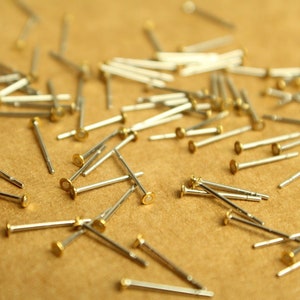 May include: A close-up of a pile of small, silver-colored metal pins with gold-colored heads. The pins are scattered on a brown surface.