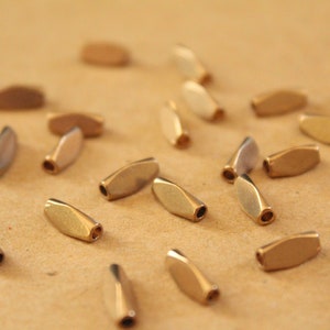May include: A collection of gold-toned metal beads, each shaped like a small oval. The beads are scattered on a light brown surface.