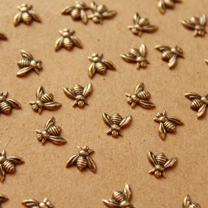 May include: A collection of gold-toned metal bee charms, scattered on a brown surface. The bees are detailed with raised lines and have a slightly textured finish.