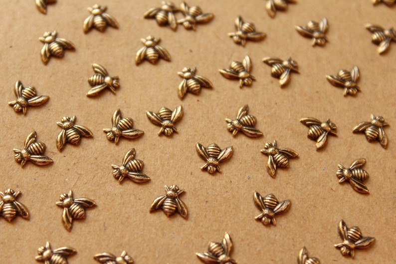 May include: A collection of gold-toned metal bee charms, scattered on a brown surface. The bees are detailed with raised lines and have a slightly textured finish.