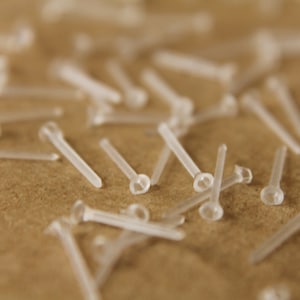 May include: A close-up of a pile of small, clear plastic push pins. The pins are scattered on a brown surface.