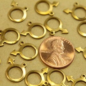 May include: A close-up of a group of gold-colored metal charms shaped like the male symbol. The charms are arranged on a brown surface with a penny in the center.