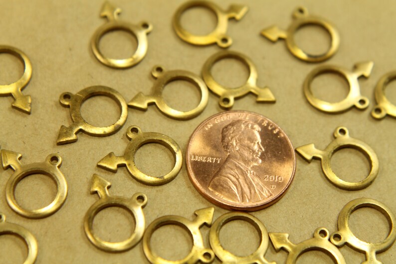 May include: A close-up of a group of gold-colored metal charms shaped like the male symbol. The charms are arranged on a brown surface with a penny in the center.