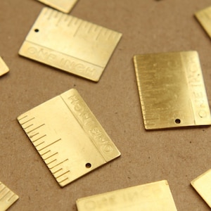 May include: A collection of eight brass ruler charms, each measuring one inch. The charms are rectangular with rounded corners and feature a ruler scale and the text "ONE INCH" engraved on each charm.