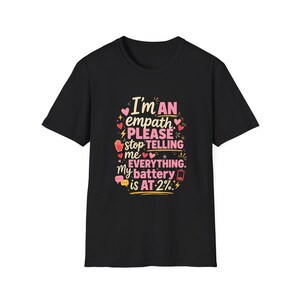 May include: A black short-sleeve t-shirt featuring colorful text that reads: I'm AN empath PLEASE stop TELLING me EVERYTHING. My battery is AT 2%. The design includes small pink hearts, yellow lightning bolts, a red hand icon, and a battery symbol.