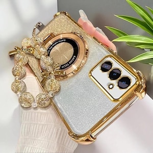 May include: A glittery silver phone case with a gold-toned frame and a circular magnetic ring mount labeled KATHY TECH. The case features a decorative wrist strap made of clear and gold-flecked spherical beads with a small gold bow charm attached.