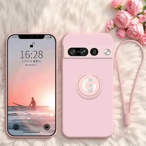 May include: A light pink smartphone case featuring a circular ring holder with a silver letter G in the center. The case includes a matching pink wrist strap and a camera cutout. The phone screen displays the time 16:28 and a sunset beach scene.
