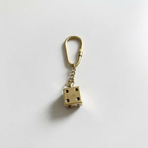 May include: A gold-coloured metal keyring featuring a single six-sided die charm attached to a small chain and a teardrop-shaped ring. The die has black circular pips on its faces, and the entire assembly is constructed from a polished metallic material.