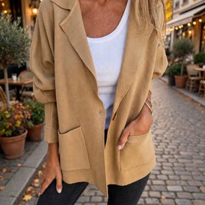 May include: A tan open-front blazer with long sleeves, a notched collar, and two front patch pockets, worn over a white ribbed tank top and dark gray jeans. The blazer features a single button closure detail and a soft, smooth texture.