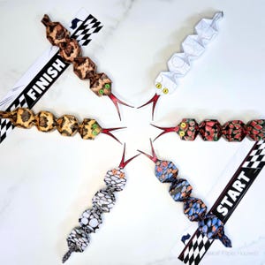 Peut inclure: Cinq serpents en papier segmentés avec des langues fourchues rouges disposés en étoile. Chaque serpent présente un motif unique incluant des écailles brunes, du blanc, du rouge et vert, du noir et blanc, et du bleu et orange. Deux lignes d'arrivée.