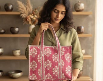 Puede incluir: Un bolso tote rectangular de tela acolchada en un tono rosa vibrante, con un patrón repetitivo de estampado floral blanco. El bolso incluye dos asas de tela a juego y una base estructurada con costuras verticales visibles en el diseño.