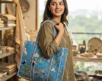 Puede incluir: Un bolso tote azul acolchado con un patrón repetitivo de cebras, tucanes y hojas de palmera tropicales. El bolso tiene dos correas de hombro resistentes con un borde a rayas blancas y azules en los bordes y las asas.
