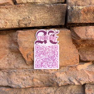 Puede incluir: Una pegatina rectangular que presenta un código QR rosa en la parte inferior y el texto "AE crafting" en color morado oscuro con una fuente gótica estilizada en la parte superior. La pegatina tiene un borde blanco y está sobre una pared de piedra.
