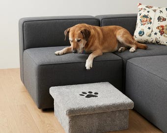 May include: A square, light gray pet step stool covered in a soft, plush fabric. The top surface features a dark gray embroidered paw print design centered on the cushion. The stool has a boxy, structured shape with a flat top and vertical sides.