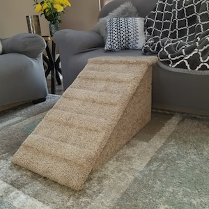 Puede incluir: Una rampa beige para mascotas con una superficie texturizada, diseñada para ayudar a las mascotas a subir y bajar de los muebles. La rampe se coloca delante de un sofá gris en un salón.