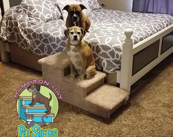 May include: Two dogs, a brown and white boxer and a brown and white mixed breed dog, are sitting on a three-step pet stairs. The stairs are covered in beige carpet. The dogs are sitting on the top two steps. The stairs are in front of a white bed with a gray and white patterned comforter. The bed is in a room with a brown carpet.
