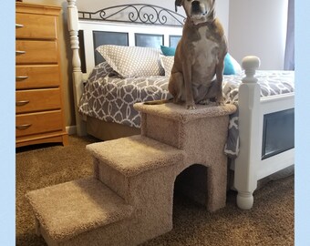 May include: Beige pet steps with a brown dog sitting on the top step. The steps have a carpeted surface and a cutout in the top step. The steps are in front of a white bed with a gray and white patterned comforter. The text "Hampton Bay Pet Steps" is visible at the bottom of the image.