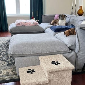 May include: A two-step pet staircase covered in beige, textured fabric. Each step features a black embroidered paw print design. The staircase is positioned in front of a large gray sectional sofa with two dogs resting on the cushions.