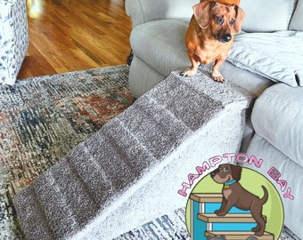 May include: A grey pet ramp with a textured surface. A brown dachshund dog is standing on the top of the ramp. The ramp is in front of a grey sofa. The image includes the logo "Hampton Bay Pet Steps".