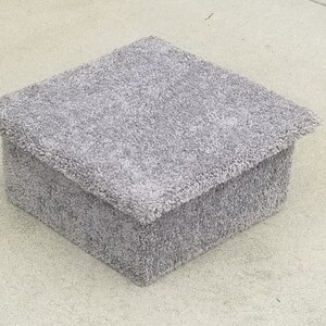 May include: A square, low-profile box covered in a thick, light gray carpet material. The box features a slightly overhanging lid with a matching textured, fuzzy surface that covers the entire exterior, creating a uniform and soft appearance.