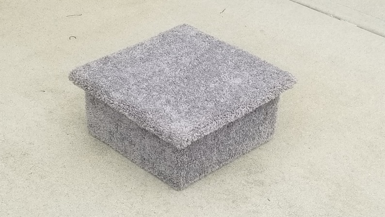 May include: A square, low-profile box covered in a thick, light gray carpet material. The box features a slightly overhanging lid with a matching textured, fuzzy surface that covers the entire exterior, creating a uniform and soft appearance.