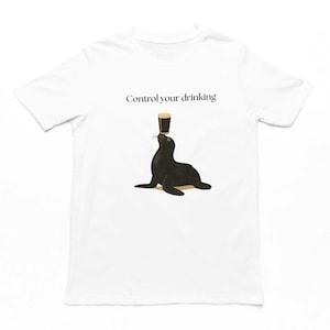 May include: A white short-sleeved t-shirt featuring a graphic of a black seal balancing a dark glass of beer on its nose. Above the seal, the text "Control your drinking" is printed in a simple black serif font. The design is centred on the chest.